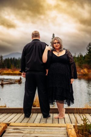 Couple stands together on wooden dock overlooking mountain lake during golden hour.