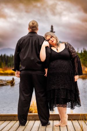 Couple shares romantic moment on lakeside dock with dramatic cloudy sky and mountains.