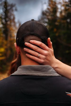 Hand with engagement ring reaches around to embrace partner wearing baseball cap.