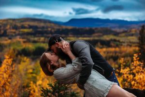 Two people share an intimate moment while standing in a field with golden fall colors and mountains behind them.