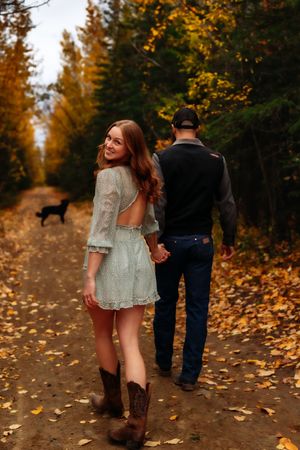 Woman in white dress walks with partner on autumn trail with dog in background.