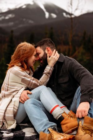 Romantic mountain engagement photo with couple in casual wear sitting on blanket.