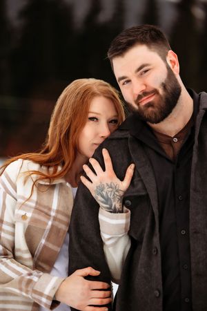 Happy couple embraces outdoors as woman shows off ring in romantic winter engagement photo.