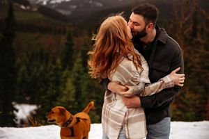 Couple embraces in snowy mountain landscape with their dog in cozy winter clothing.