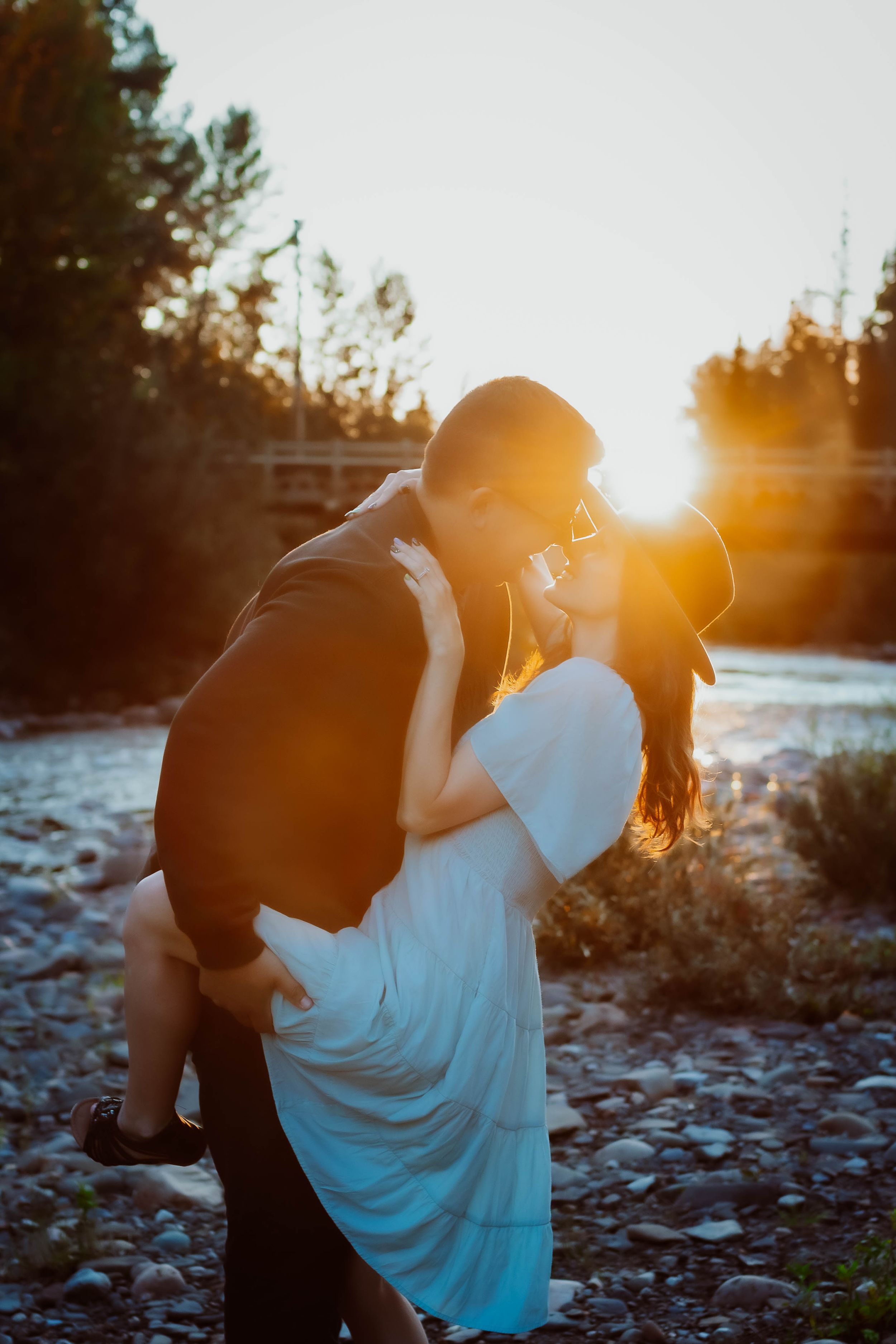 Romantic sunset couple embracing by river with sunburst effect through trees.