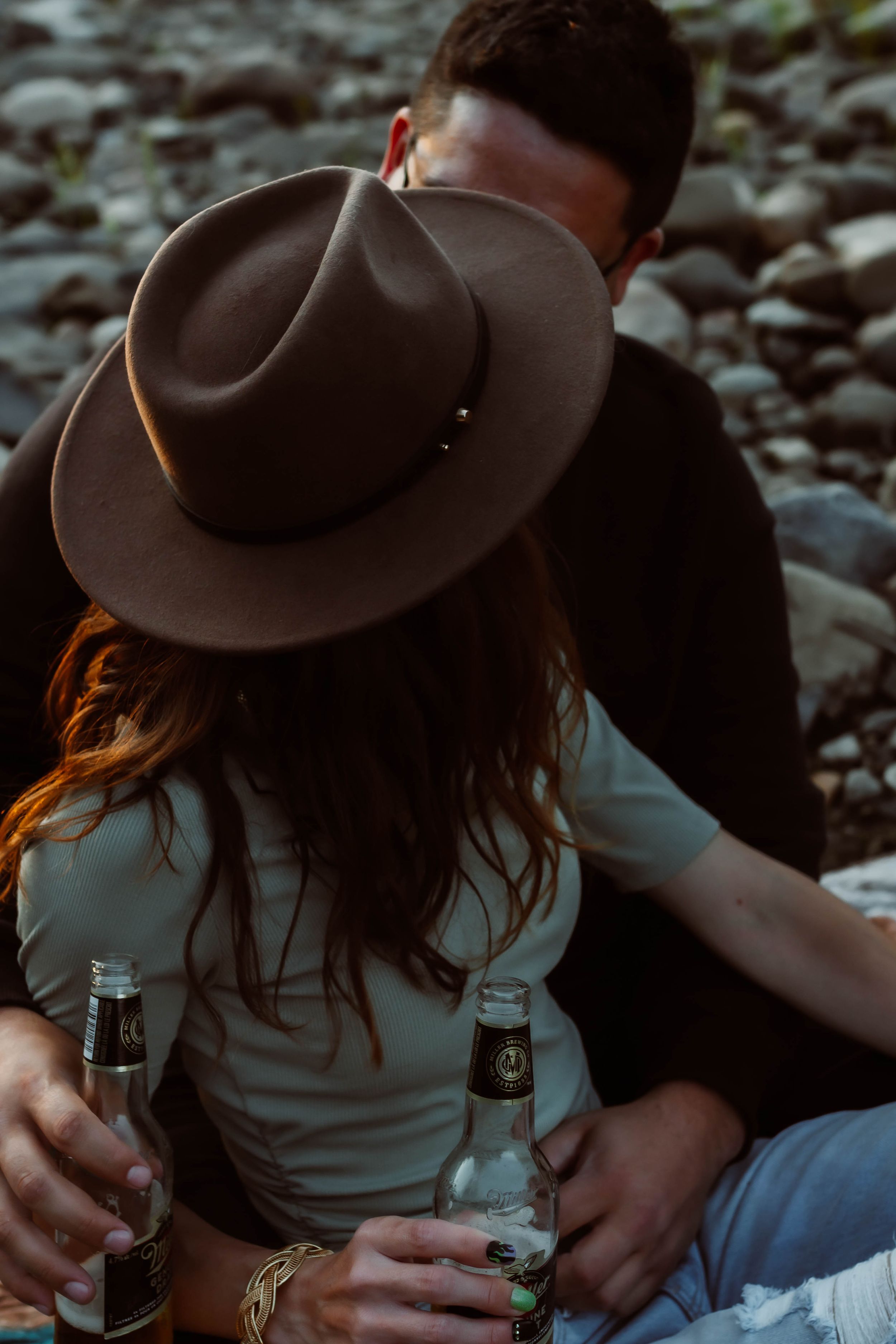 Couple sharing drinks on rocky shore with woman wearing brown fedora hat.