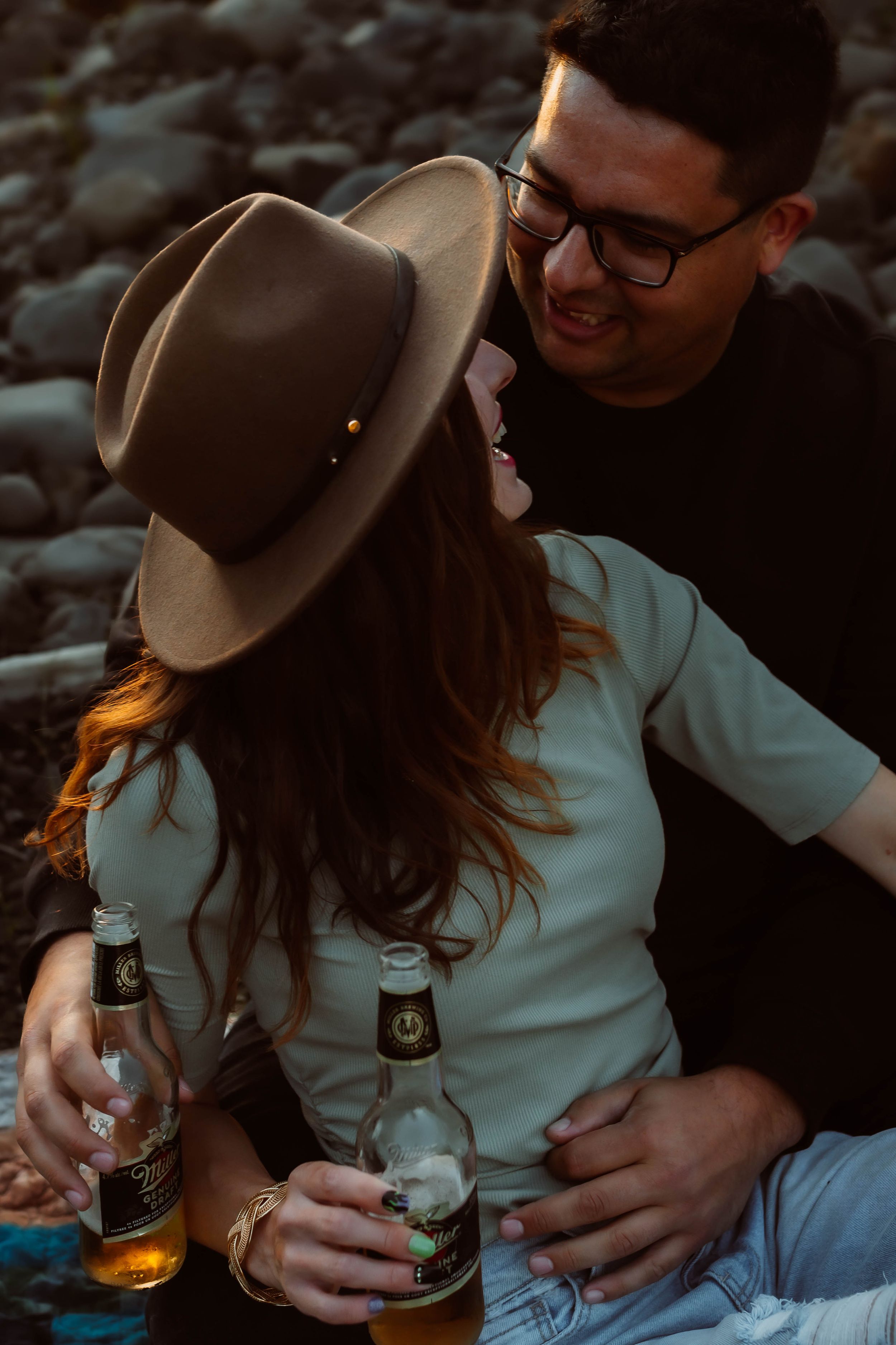 Intimate moment between couple sharing drinks on rocky shore at sunset.