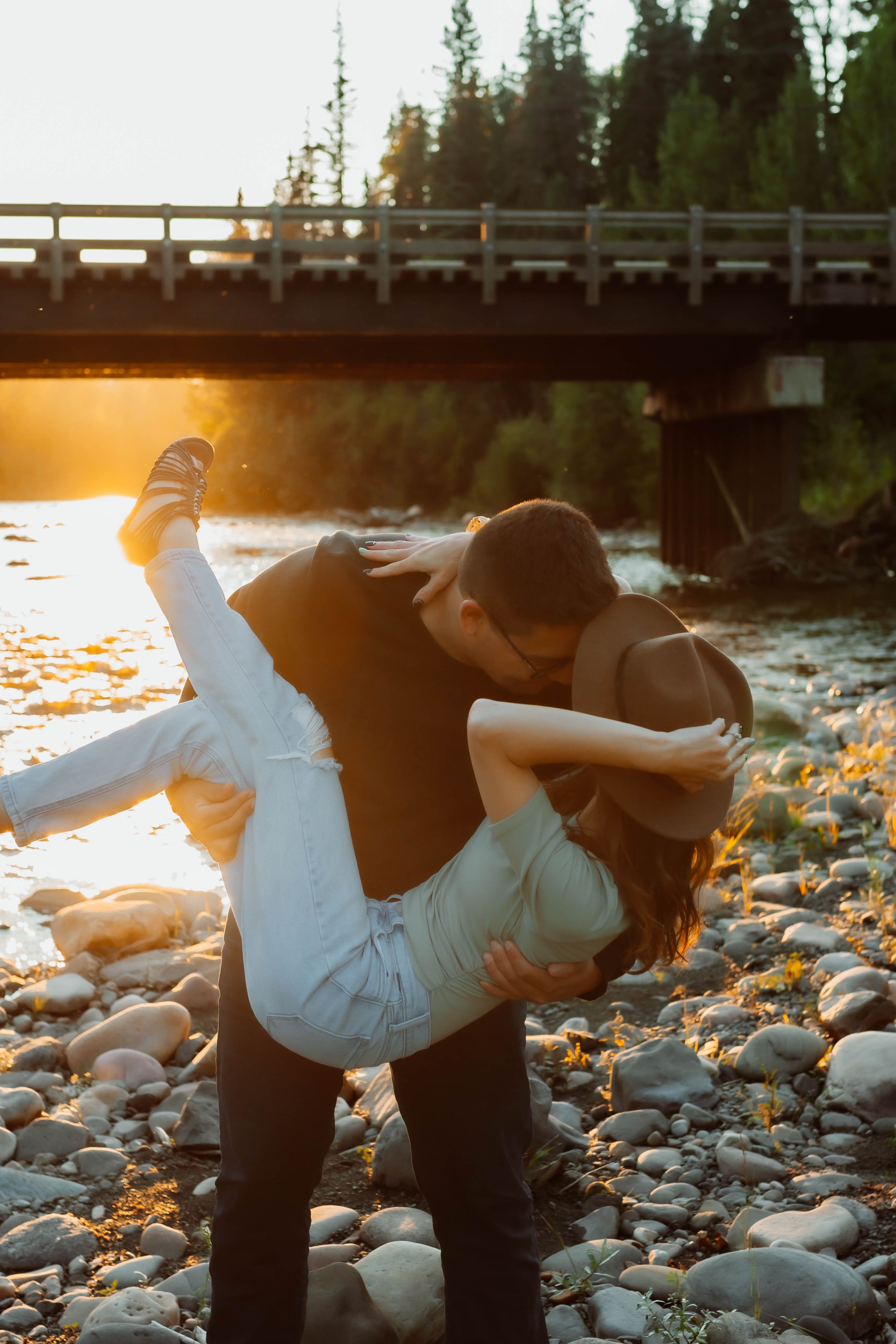 Playful couple dancing on rocky riverbank during golden hour with bridge in background.