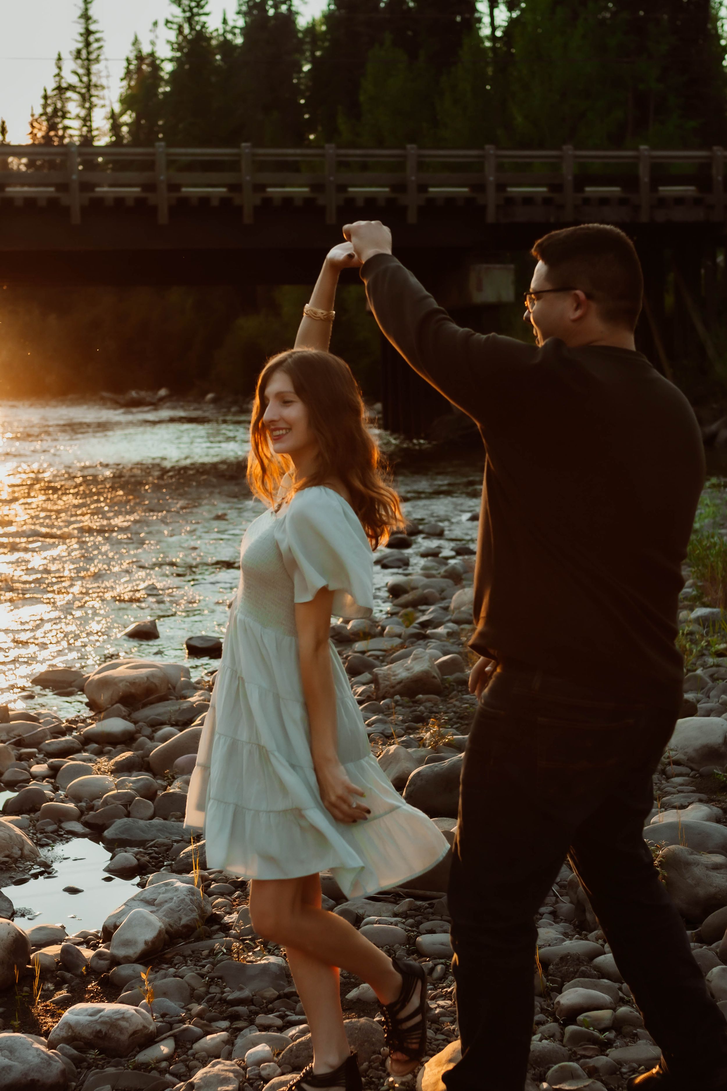 Couple dancing on rocky riverbank during golden hour with bridge silhouette in background.