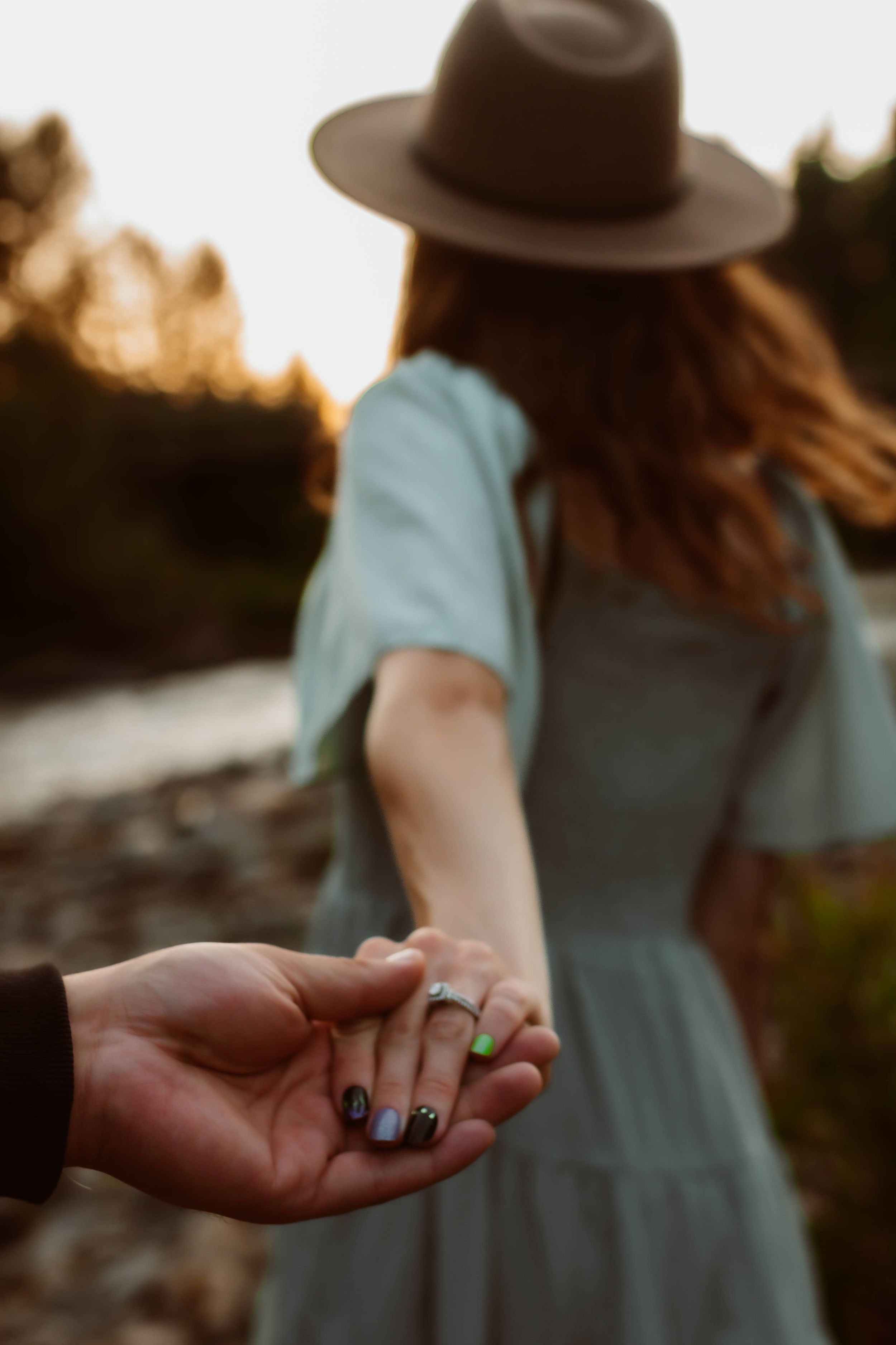 Person in blue dress and brown hat holds hands while showing engagement ring.