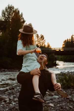 Playful couple having fun by river with person in hat sitting on partner's shoulders.