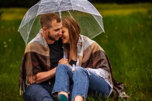 Couple snuggling under clear umbrella in rainy field with blanket wrapped around them.