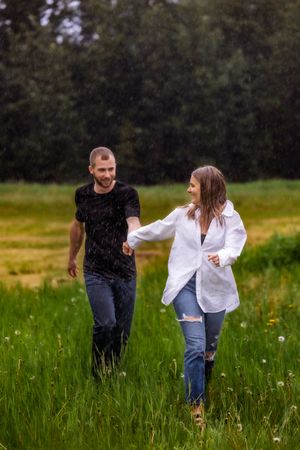 Couple running playfully through tall grass during summer rain shower.