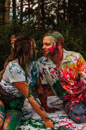 Two people covered in vibrant paint having a playful moment in a forest setting.