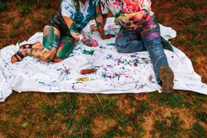 Paint-splattered white sheet on grass with couple sitting during colorful photoshoot.