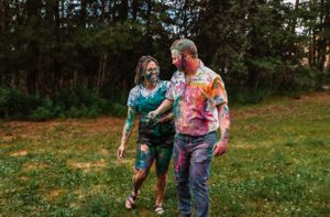 Paint-covered couple walks together through green grass during artistic photoshoot.