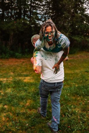 Playful piggyback ride during outdoor paint photoshoot in grassy field.