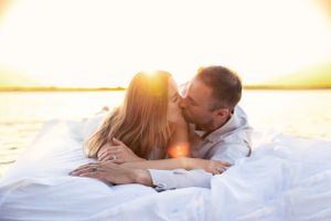 Couple sharing intimate moment while lying on white blanket at sunset by water.