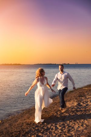 A couple runs playfully along a beach shoreline during a golden sunset.