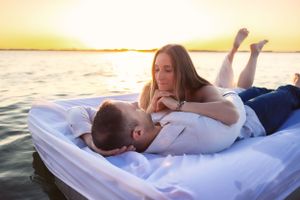 A couple relaxes on white blankets during a beachside sunset photoshoot.