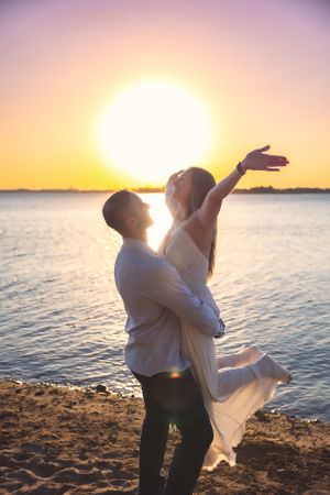 Couple dancing joyfully on beach at sunset with woman in flowing dress being lifted.