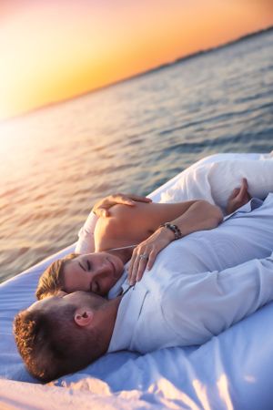 Couple lying together on white blanket by water during sunset.