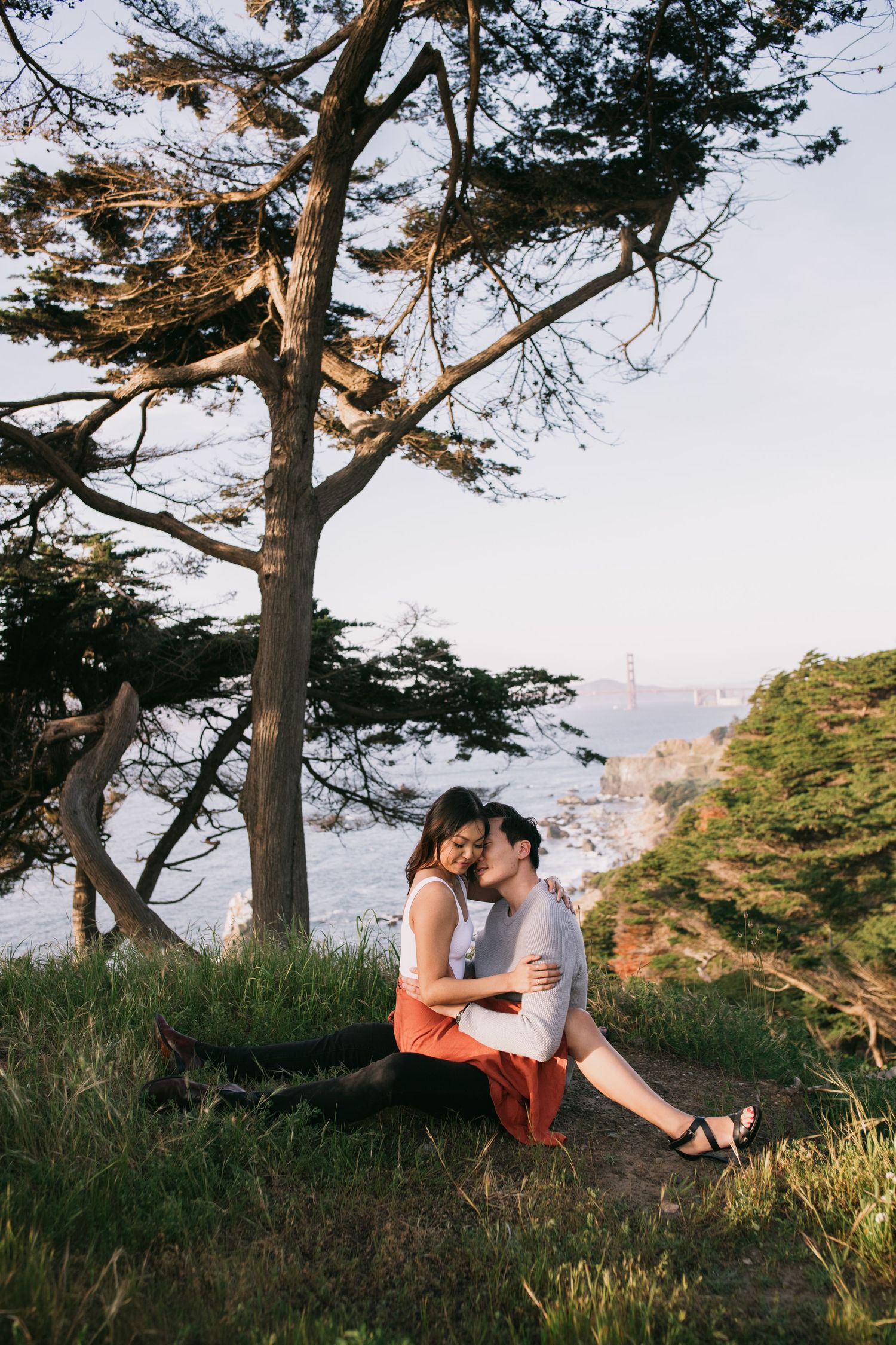 Couple shares a tender kiss on a coastal stone wall with crashing ocean waves behind them.