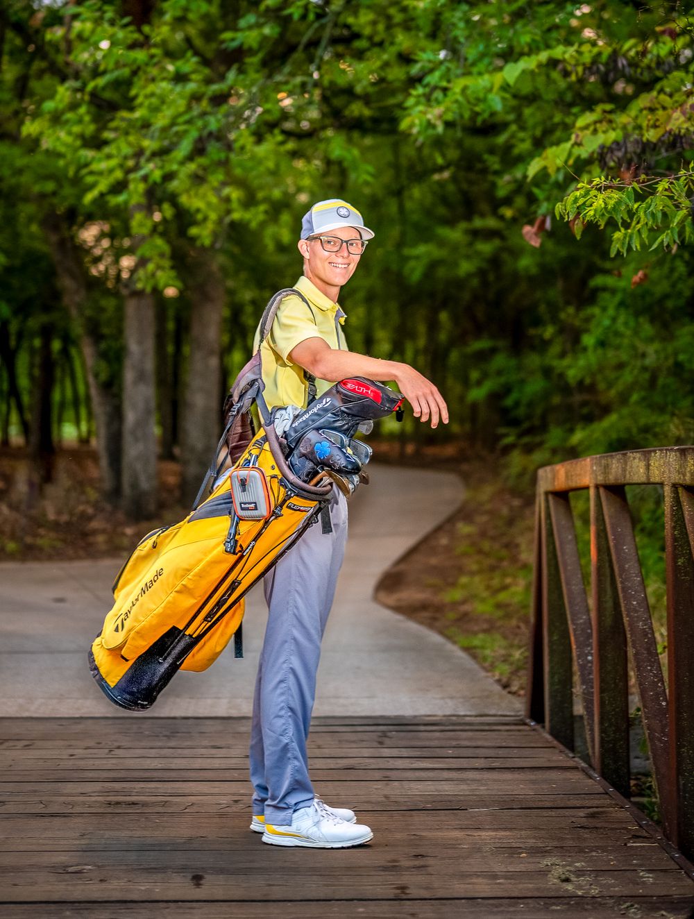 A golfer stands on a wooden bridge carrying a yellow golf bag on a tree-lined path in summer.