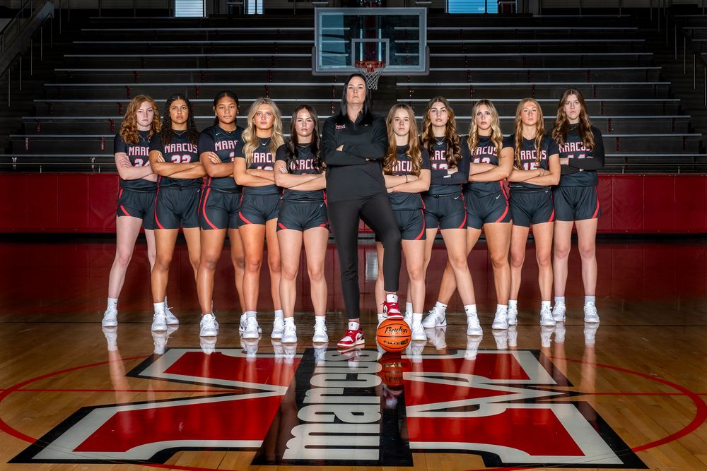 Athletic team poses together on wooden basketball court with school logo, wearing matching navy uniforms.