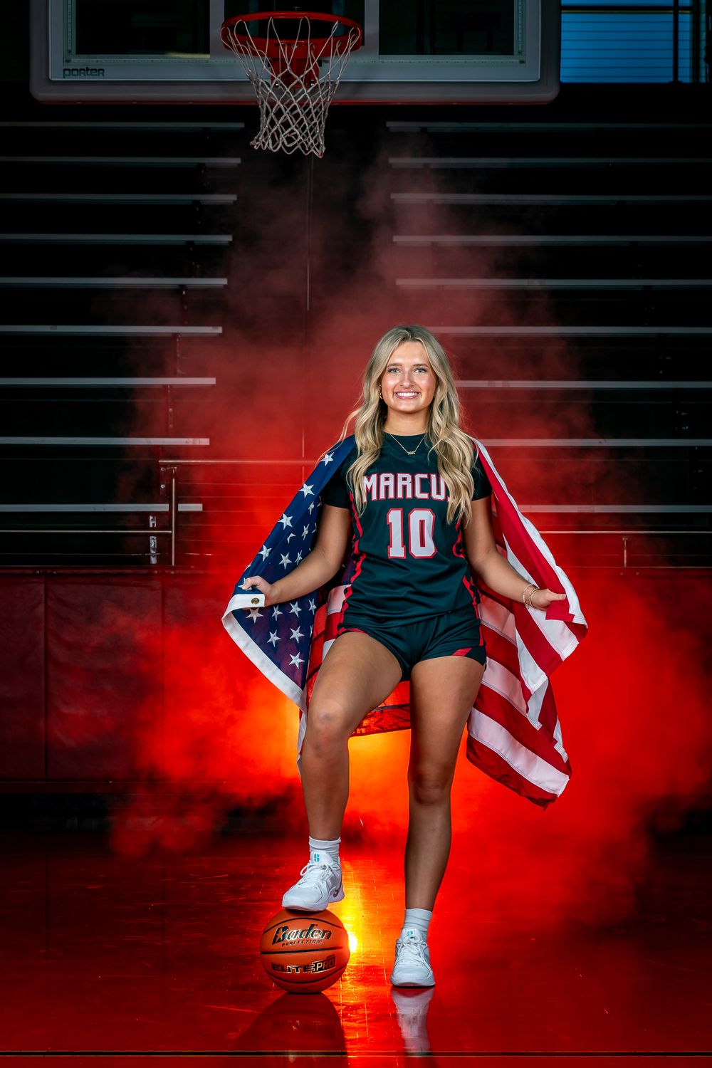 Basketball player poses in dark uniform with number 10 and American flag against red smoke on indoor court.