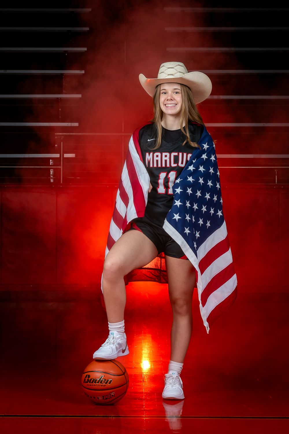 Basketball player poses with American flag and cowboy hat in front of red backdrop with basketball at their feet.
