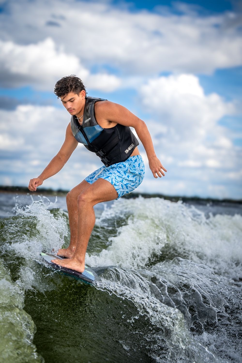 A wakeboarder in blue swim shorts and black life vest rides a wave on a lake under cloudy summer skies.