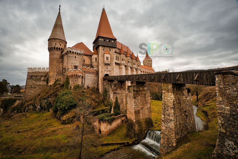 Medieval Corvin Castle in Romania perched on a hill with dramatic cloudy sky backdrop.