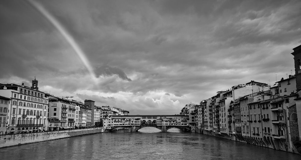 Black and white photo of Ponte Vecchio bridge over Arno River in Florence, Italy, with a rainbow in dramatic cloudy sky.