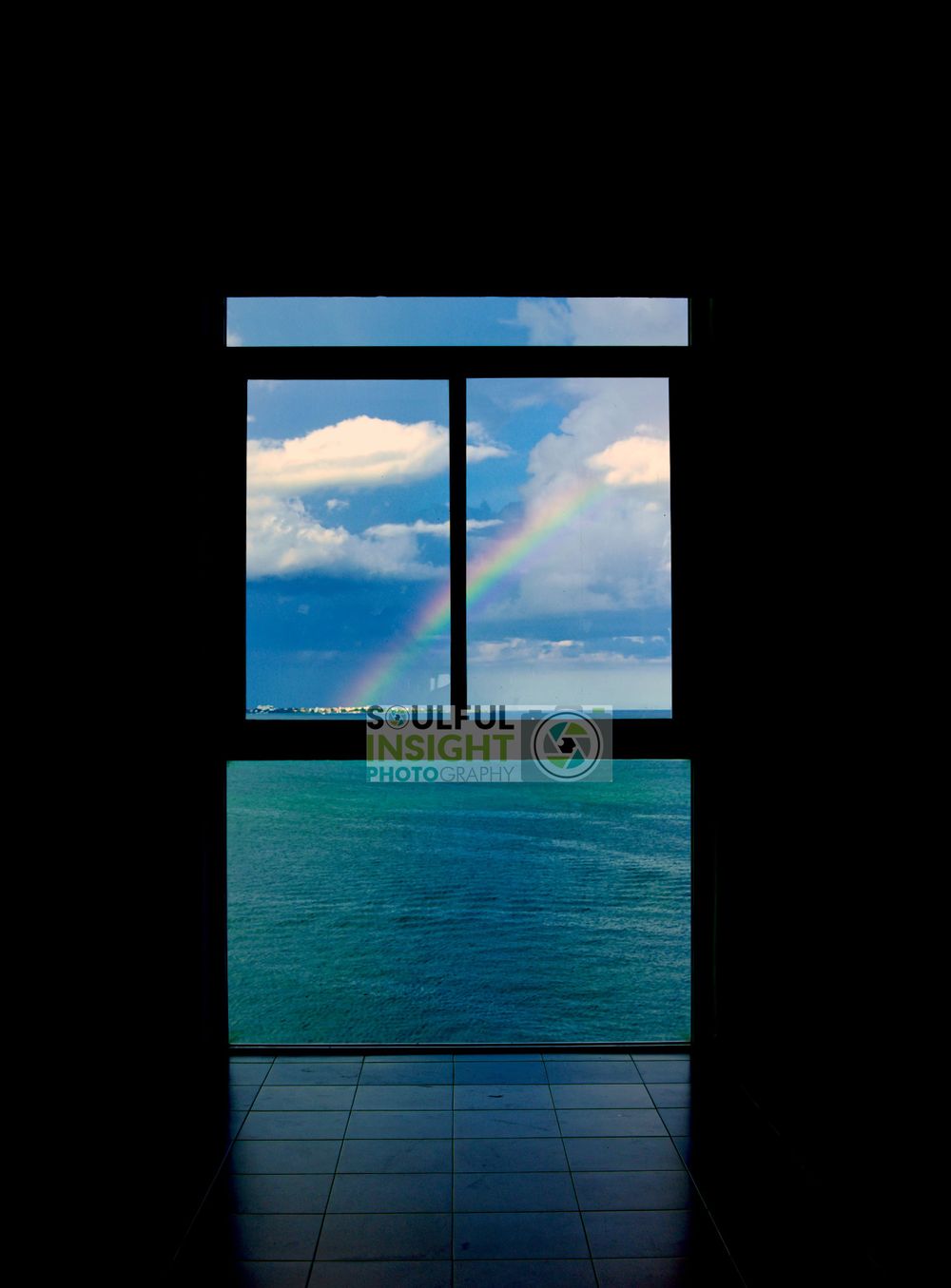A rainbow appears over turquoise waters viewed through a dark window frame.