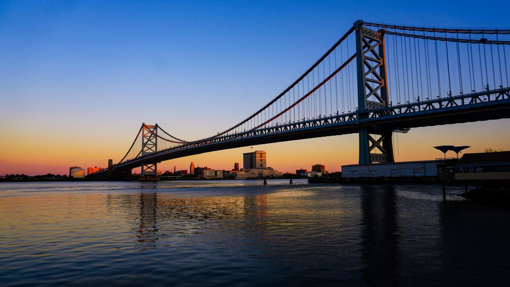 Ben Franklin Bridge spans the Delaware River at sunset with Philadelphia skyline in silhouette.