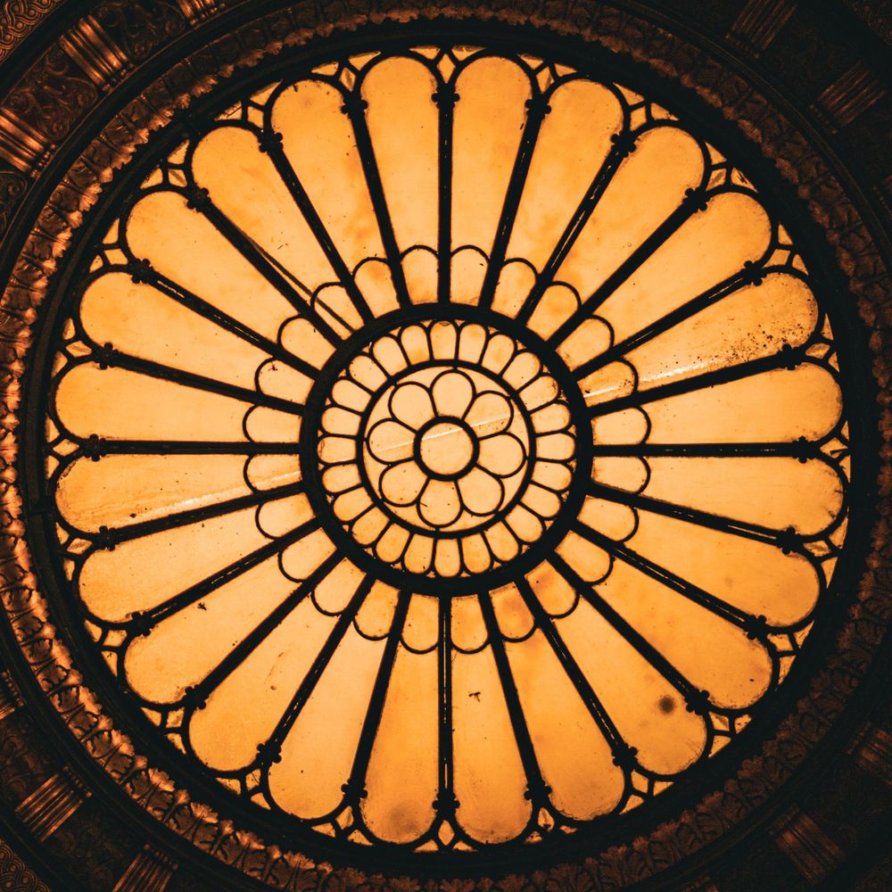 Ornate circular stained glass window with orange light illuminating an intricate floral rosette pattern.
