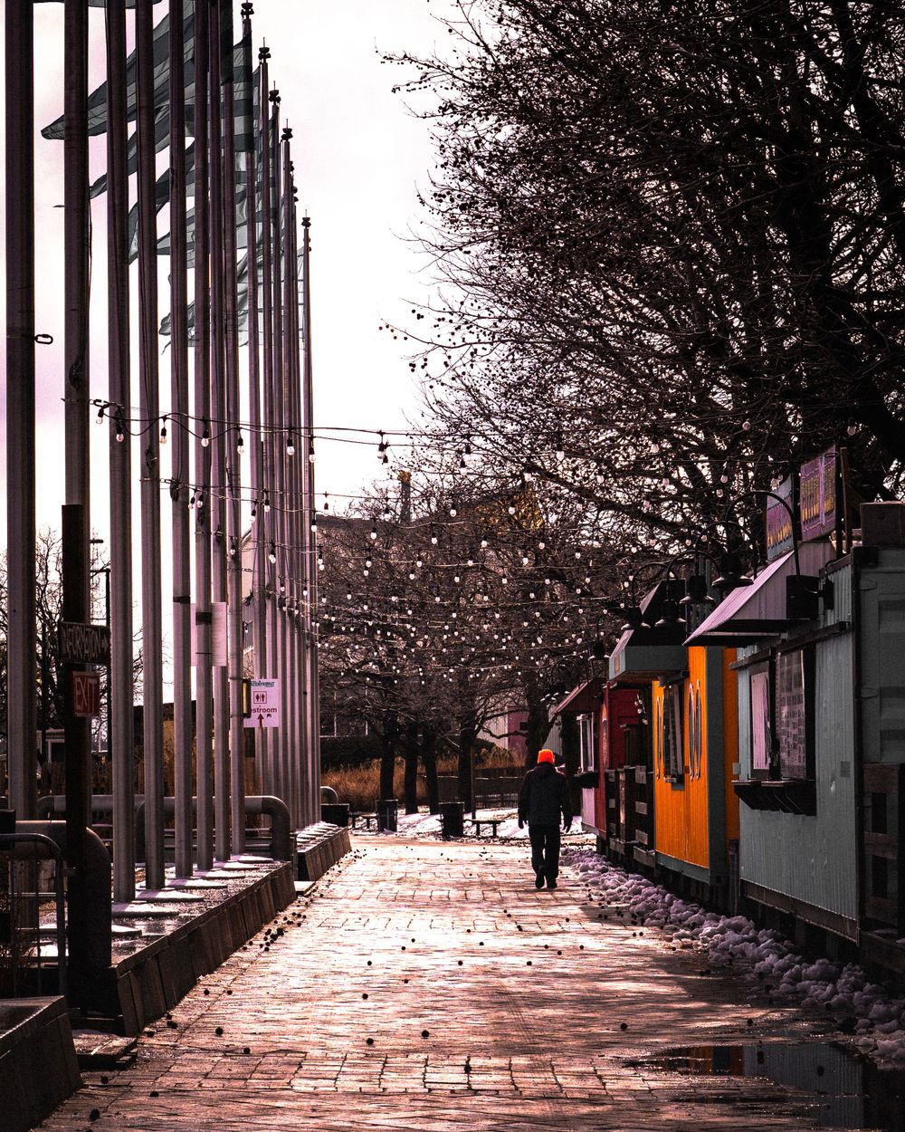 Urban street scene with tall metal pillars and snow-covered sidewalk leading to colorful buildings.
