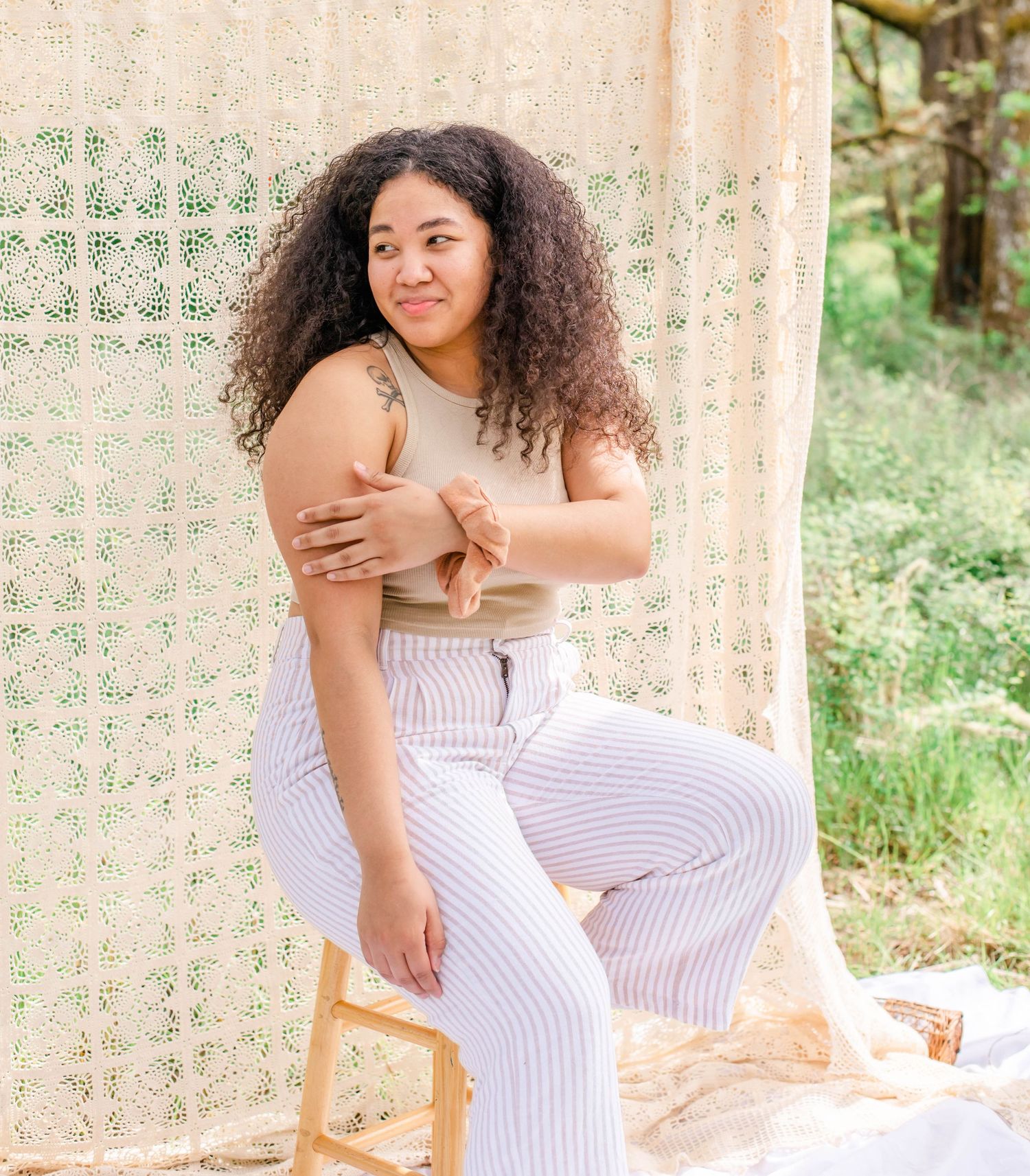 Natural light photography setup with macrame backdrop and simple wooden chair against green foliage.
