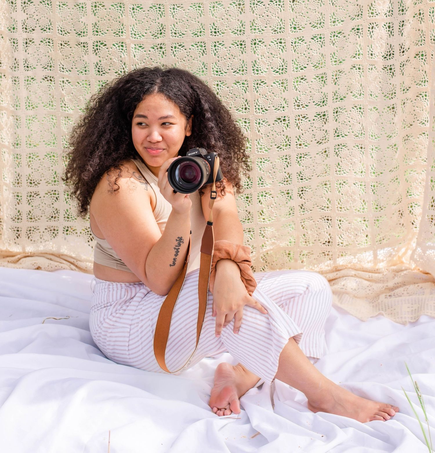 Photographer with curly hair lying on white sheets holds camera in casual boudoir photoshoot setting.