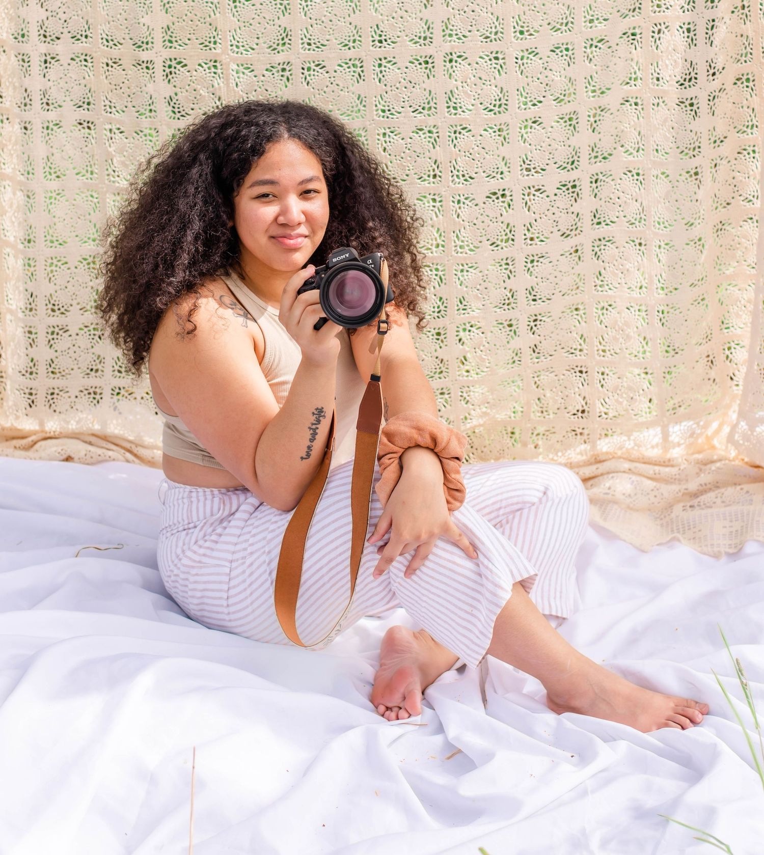 Photographer with curly hair lying on white sheets holds camera in casual boudoir photoshoot setting.