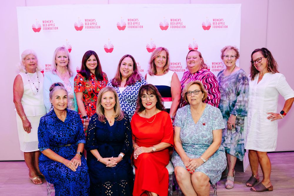 Group photo of women in colorful dresses at foundation event with pink backdrop.