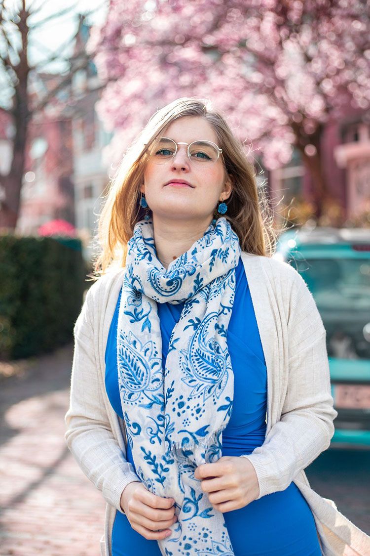 A woman in a blue top and white cardigan poses with a blue and white patterned scarf under blooming magnolia blossoms.
