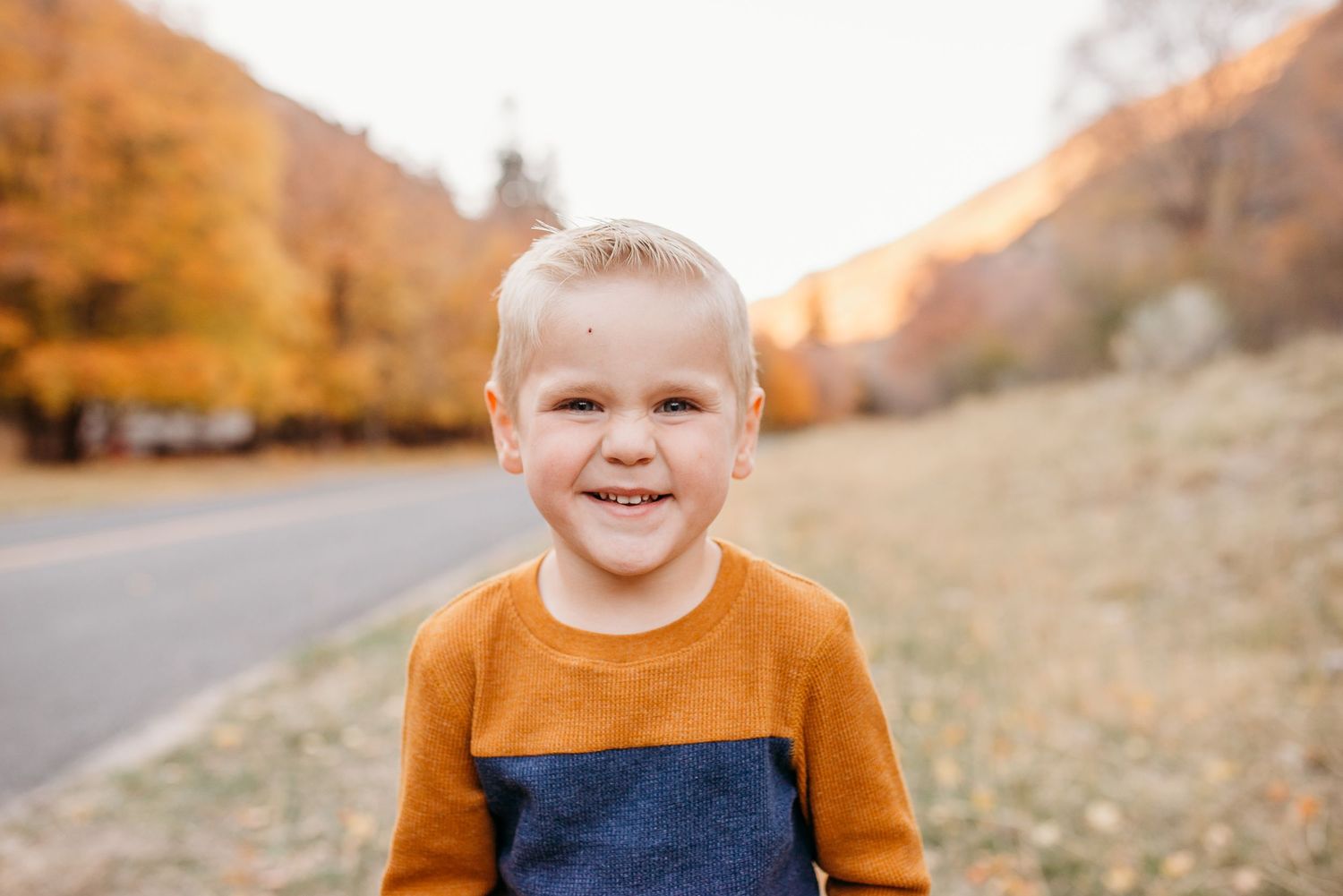 A small child wearing an orange and navy sweater smiles brightly on an autumn mountain road.