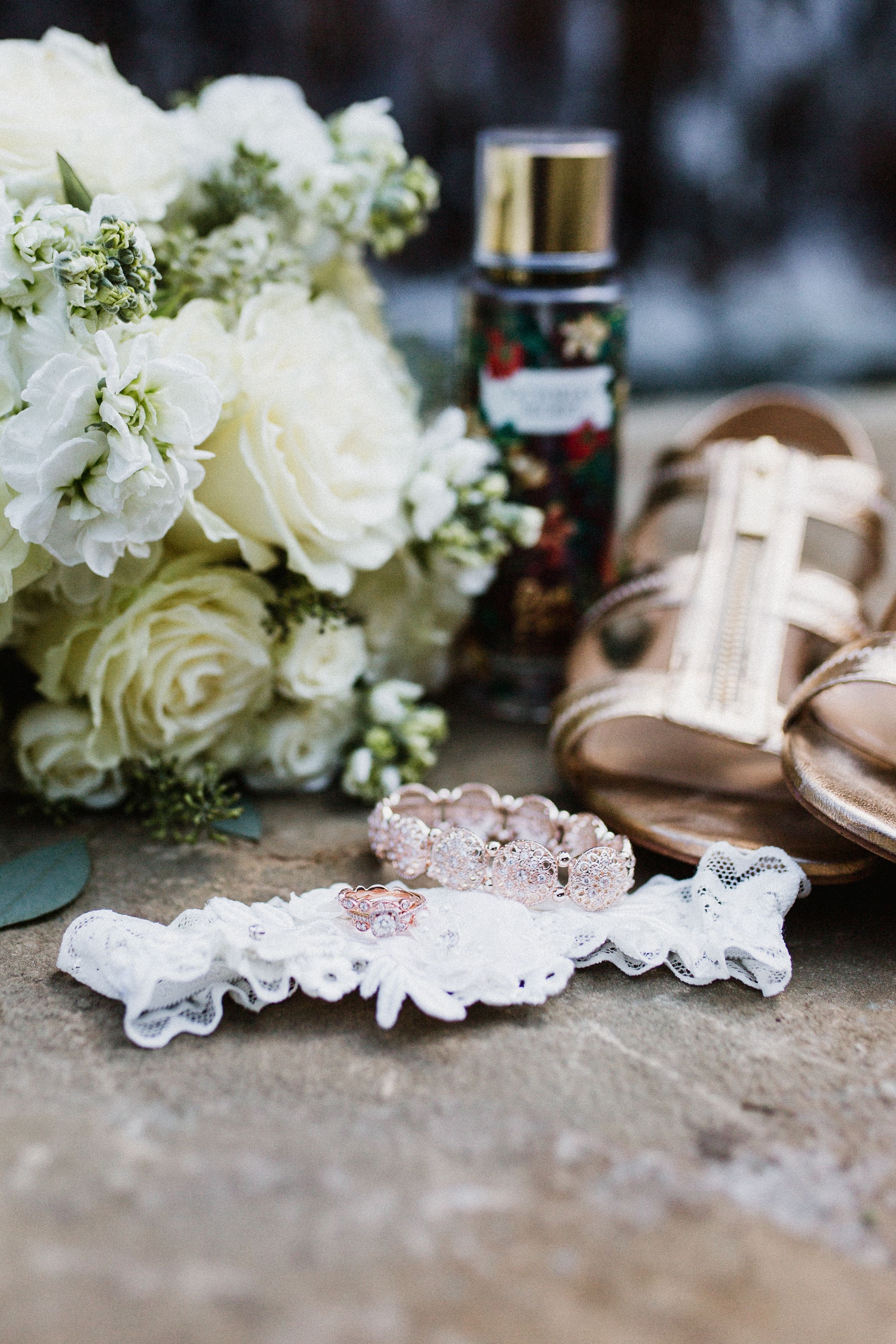 Wedding accessories including a white rose bouquet, lace garter, perfume bottle and blush colored shoes arranged on stone surface.