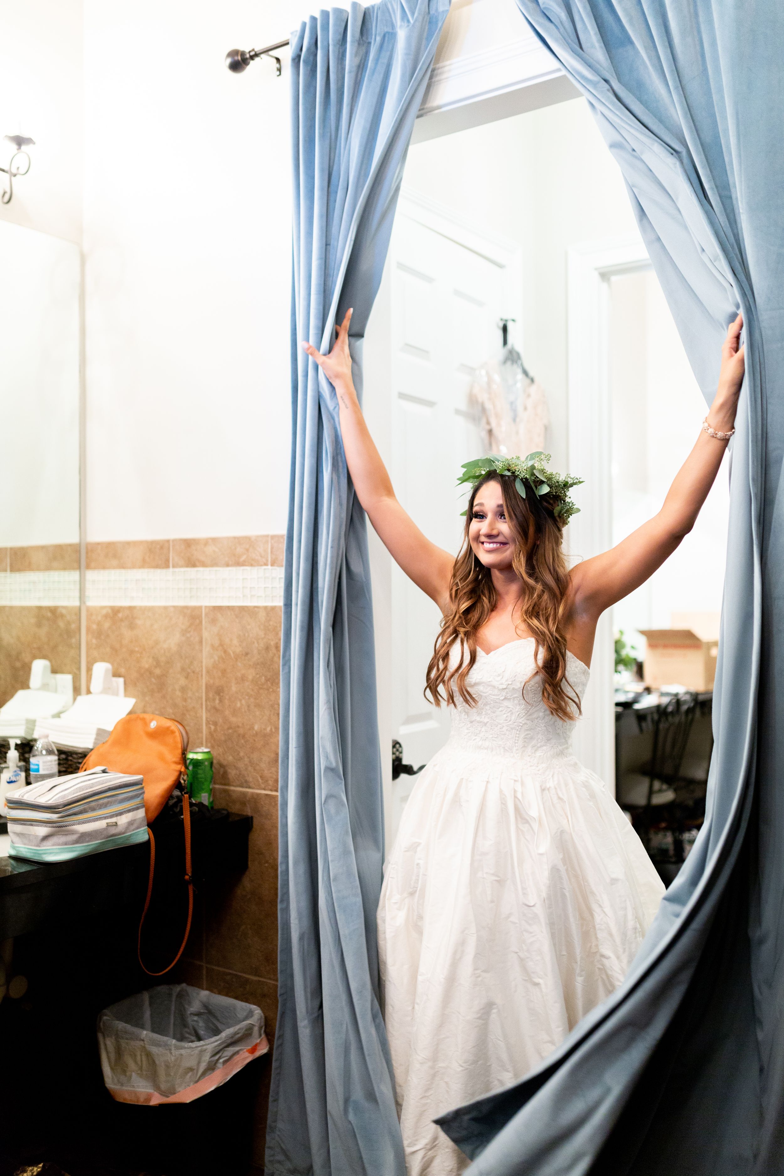 A bride in a white wedding dress joyfully parts blue curtains in a getting ready room.