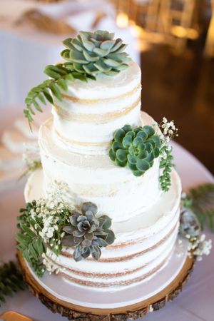 Semi-naked white wedding cake decorated with fresh succulents and greenery on a wooden cake stand.