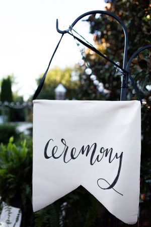 White fabric banner with calligraphy reading 'ceremony' hanging on black metal stand.