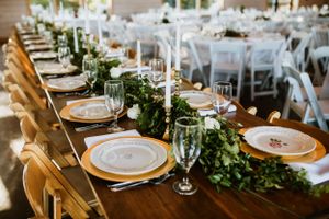 Long wooden farm table set for wedding reception with garland runner and gold charger plates.