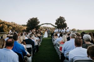 Outdoor wedding ceremony setup with white chairs on grassy hillside under archway.